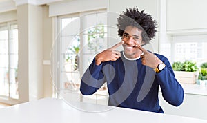 Young african american man wearing casual sweater sitting at home Smiling with open mouth, fingers pointing and forcing cheerful