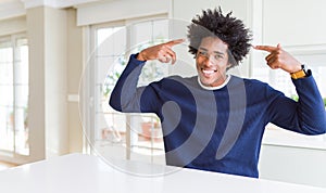 Young african american man wearing casual sweater sitting at home smiling pointing to head with both hands finger, great idea or