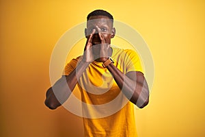 Young african american man wearing casual t-shirt standing over isolated yellow background Shouting angry out loud with hands over