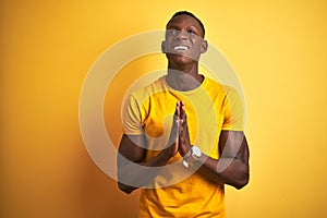 Young african american man wearing casual t-shirt standing over  yellow background begging and praying with hands together