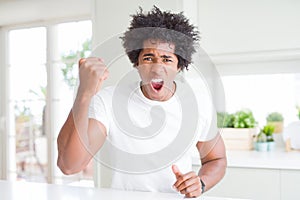 Young african american man wearing casual white t-shirt sitting at home angry and mad raising fist frustrated and furious while