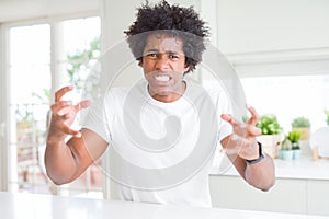 Young african american man wearing casual white t-shirt sitting at home Shouting frustrated with rage, hands trying to strangle,