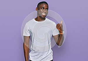 Young african american man wearing casual white t shirt smiling with happy face looking and pointing to the side with thumb up