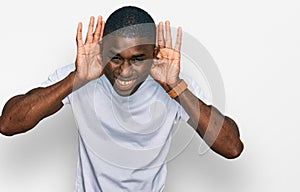 Young african american man wearing casual white t shirt trying to hear both hands on ear gesture, curious for gossip