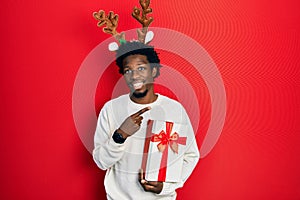 Young african american man wearing deer christmas hat holding gift cheerful with a smile on face pointing with hand and finger up