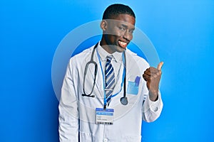 Young african american man wearing doctor uniform smiling with happy face looking and pointing to the side with thumb up