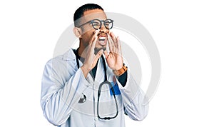 Young african american man wearing doctor uniform and stethoscope shouting angry out loud with hands over mouth