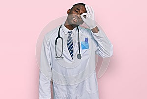 Young african american man wearing doctor uniform and stethoscope smiling happy doing ok sign with hand on eye looking through