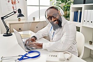 Young african american man wearing doctor uniform using laptop and headphones working at clinic