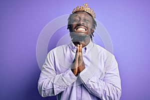 Young african american man wearing golden crown of king over isolated purple background begging and praying with hands together