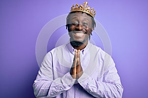 Young african american man wearing golden crown of king over isolated purple background praying with hands together asking for