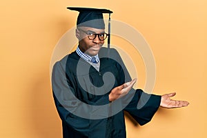 Young african american man wearing graduation cap and ceremony robe inviting to enter smiling natural with open hand