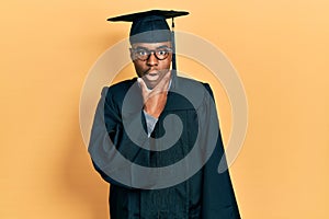 Young african american man wearing graduation cap and ceremony robe looking fascinated with disbelief, surprise and amazed