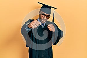 Young african american man wearing graduation cap and ceremony robe punching fist to fight, aggressive and angry attack, threat