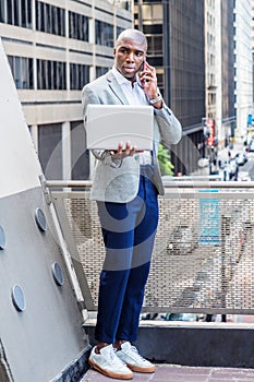 Young African American man talking on cell phone, working on laptop computer outside in New York City