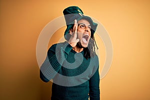 Young african american man wearing green hat with clover celebrating saint patricks day shouting and screaming loud to side with