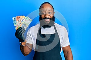 Young african american man wearing professional apron holding australia dollars banknotes looking positive and happy standing and