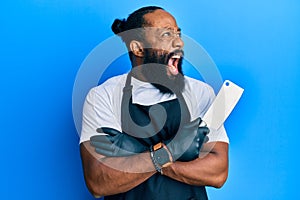 Young african american man wearing professional apron holding knife angry and mad screaming frustrated and furious, shouting with