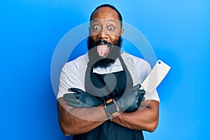 Young african american man wearing professional apron holding knife sticking tongue out happy with funny expression