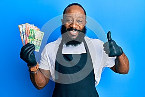 Young african american man wearing professional apron holding south africa rand banknotes smiling happy and positive, thumb up