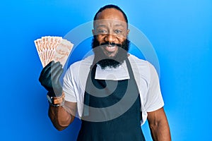 Young african american man wearing professional apron holding turkish liras banknotes looking positive and happy standing and