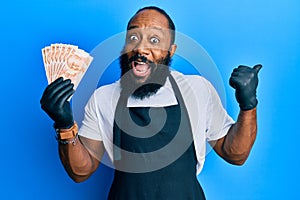 Young african american man wearing professional apron holding turkish liras banknotes pointing thumb up to the side smiling happy