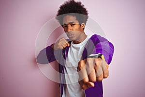 Young african american man wearing purple sweatshirt standing over isolated pink background Punching fist to fight, aggressive and
