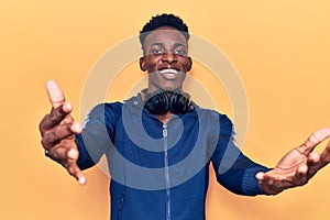 Young african american man wearing sportswear and headphones looking at the camera smiling with open arms for hug