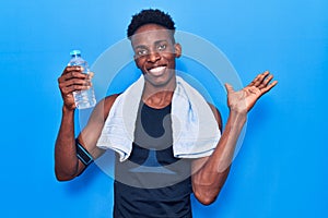 Young african american man wearing sportswear and towel holding bottle of water celebrating achievement with happy smile and