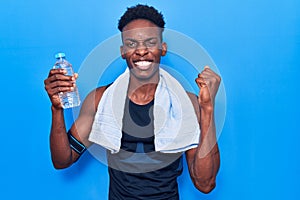 Young african american man wearing sportswear and towel holding bottle of water screaming proud, celebrating victory and success