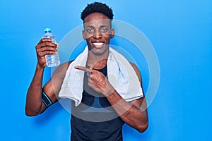 Young african american man wearing sportswear and towel holding bottle of water smiling happy pointing with hand and finger