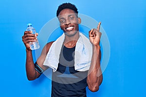 Young african american man wearing sportswear and towel holding bottle of water smiling happy pointing with hand and finger to the