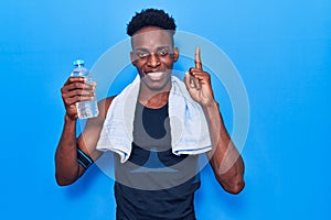 Young african american man wearing sportswear and towel holding bottle of water smiling with an idea or question pointing finger