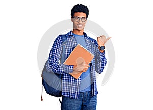 Young african american man wearing student backpack holding book pointing to the back behind with hand and thumbs up, smiling