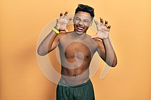 Young african american man wearing swimwear and swimmer glasses smiling funny doing claw gesture as cat, aggressive and sexy