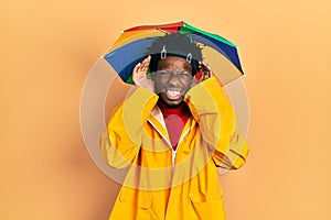 Young african american man wearing yellow raincoat trying to hear both hands on ear gesture, curious for gossip