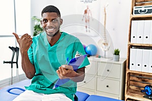 Young african american man working at pain recovery clinic pointing to the back behind with hand and thumbs up, smiling confident