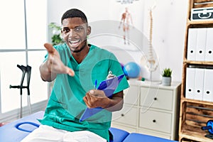 Young african american man working at pain recovery clinic smiling friendly offering handshake as greeting and welcoming