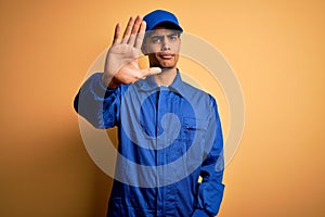 Young african american mechanic man wearing blue uniform and cap over yellow background doing stop sing with palm of the hand