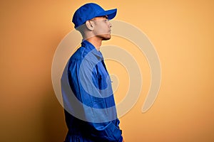 Young african american mechanic man wearing blue uniform and cap over yellow background looking to side, relax profile pose with