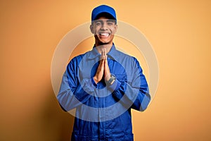 Young african american mechanic man wearing blue uniform and cap over yellow background praying with hands together asking for