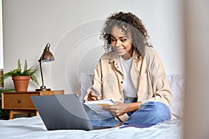 Young African American mixed race student girl sitting on bed with pc indoors.