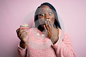 Young african american plus size woman with braids eating sweet chocolate cupcake cover mouth with hand shocked with shame for