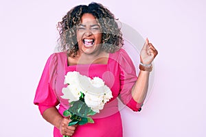 Young african american plus size woman holding flowers screaming proud, celebrating victory and success very excited with raised
