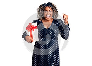 Young african american plus size woman holding gift screaming proud, celebrating victory and success very excited with raised arms