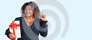Young african american plus size woman holding gift surprised with an idea or question pointing finger with happy face, number one