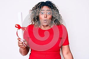 Young african american plus size woman holding graduate degree diploma scared and amazed with open mouth for surprise, disbelief