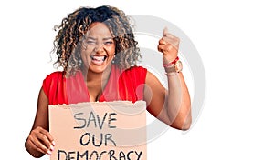 Young african american plus size woman holding save our democracy banner screaming proud, celebrating victory and success very