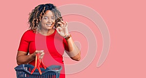 Young african american plus size woman holding supermarket shopping basket doing ok sign with fingers, smiling friendly gesturing