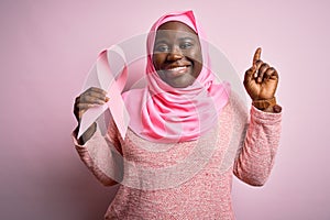 Young african american plus size woman wearing muslim hijab holding pink cancer ribbon surprised with an idea or question pointing
